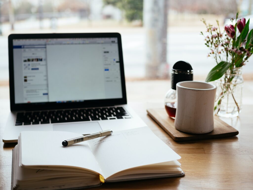 A notebook, laptop, coffee cup, and jar of flowers on a desk.