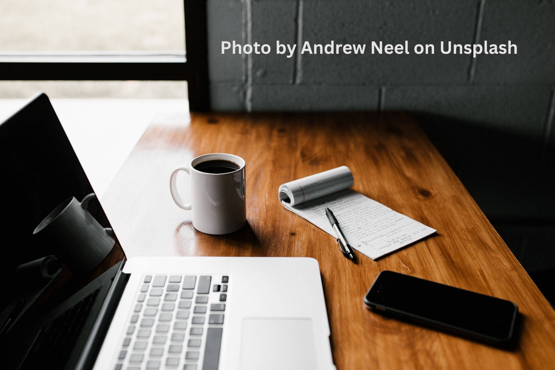 A laptop, coffee cup, cell phone, and note pad sitting on a wooden table. With the words, "Photo by Andrew Neel on Unsplash" in the corner.
